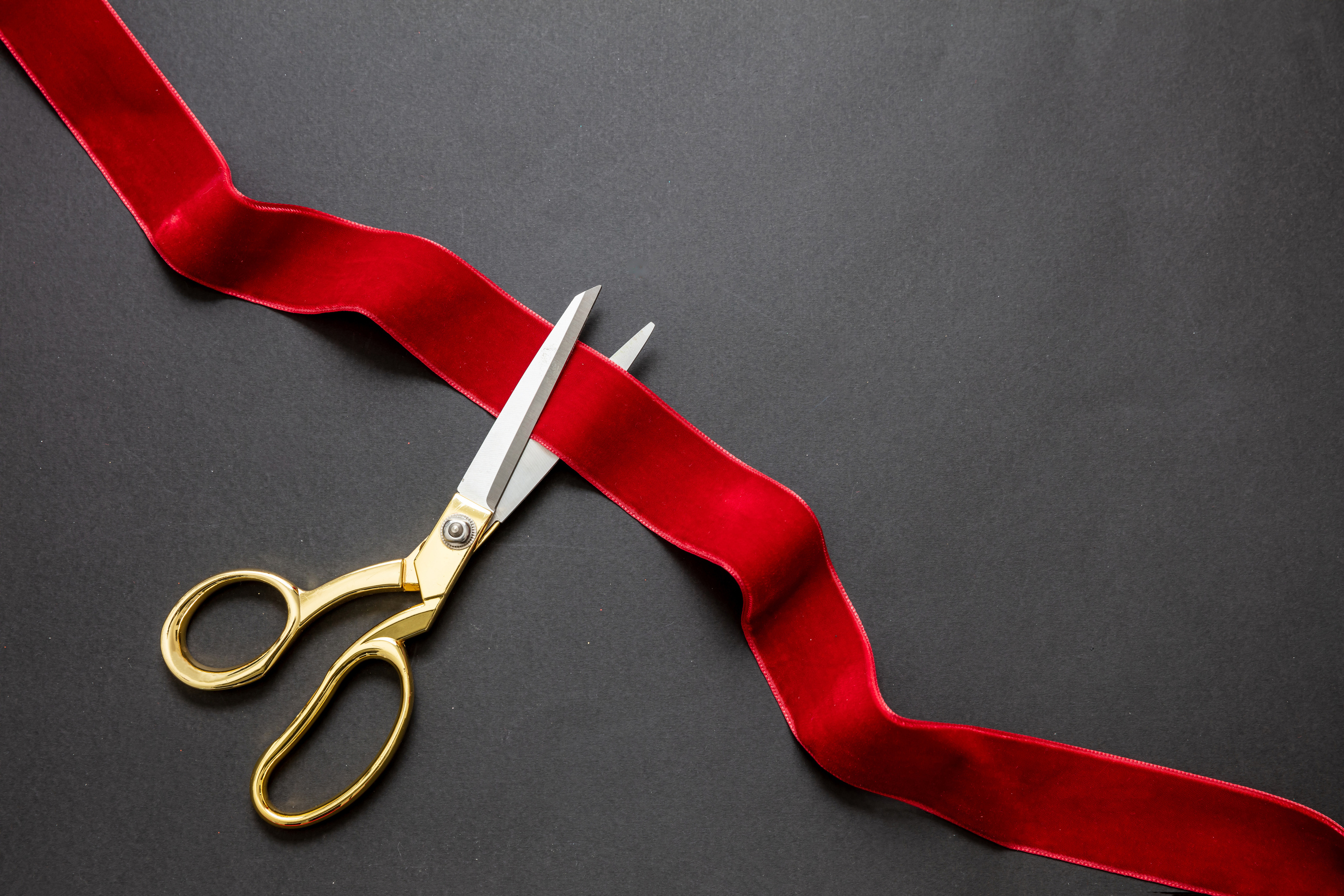 Grand opening, ribbon cut, overhead of gold scissors cutting red velvet ribbon on black